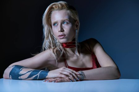 Foto de Mujer joven de moda con resaltador y tatuaje sentado y mirando hacia otro lado en el fondo oscuro - Imagen libre de derechos