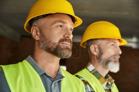 hombres barbudos atractivos en cascos de seguridad y chalecos posando y mirando hacia otro lado, constructores de cabañas