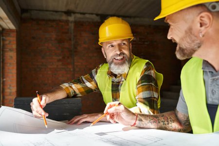 Photo for Cheerful good looking builders in safety vests and helmets looking at blueprint of building - Royalty Free Image