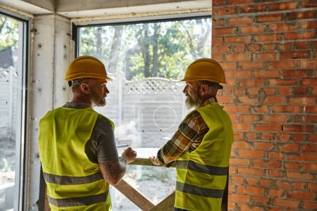 Foto de Dos hombres barbudos trabajadores en chalecos de seguridad y cascos en el sitio de construcción, constructores de cabañas - Imagen libre de derechos