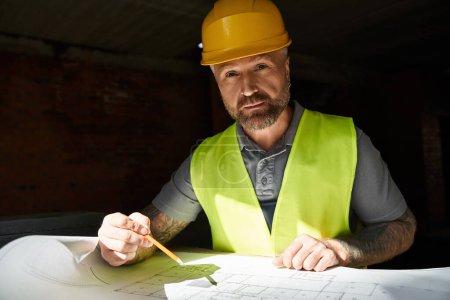 handsome builder in safety vest working on his blueprint on construction site and looking at camera