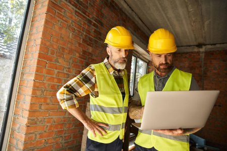 Photo for Good looking construction workers in safety vests working with laptop on site, cottage builders - Royalty Free Image