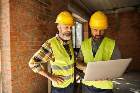Photo for Cheerful devoted construction workers in safety vests working with laptop on site, cottage builders - Royalty Free Image