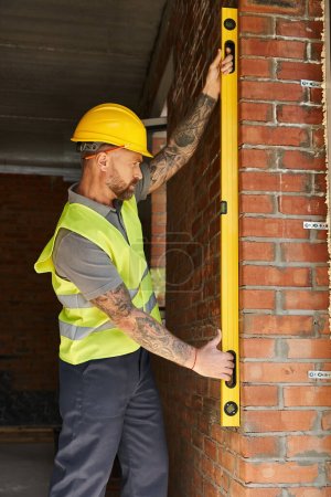 guapo barbudo trabajador de la construcción en chaleco de seguridad que mide la pared de ladrillo con nivel, constructor