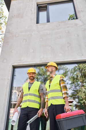 Téléchargez les photos : Travailleurs de la construction joyeux avec le niveau de la boîte à outils et le plan en regardant la caméra à l'extérieur - en image libre de droit