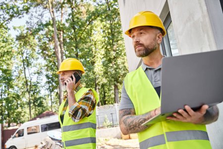 appealing builder with tattoos working on laptop while his colleague talking by phone during work