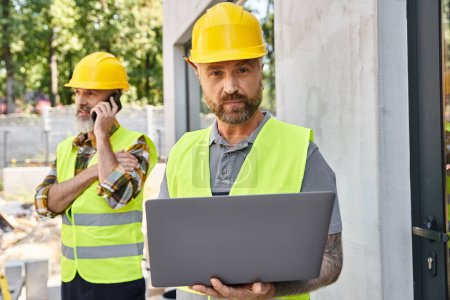 Photo for Appealing builder with tattoos working on laptop while his colleague talking by phone during work - Royalty Free Image