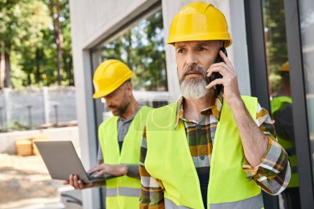 Photo for Attractive builder with tattoos working on laptop while his colleague talking by phone during work - Royalty Free Image