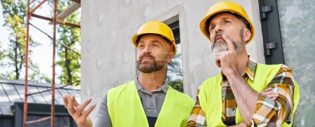 Photo for Two good looking workers in safety vests and helmets discussing site, cottage builders, banner - Royalty Free Image