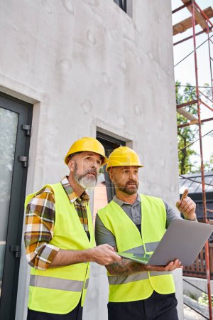 Photo for Handsome bearded men in safety vests and helmets working with laptop on construction site, builders - Royalty Free Image