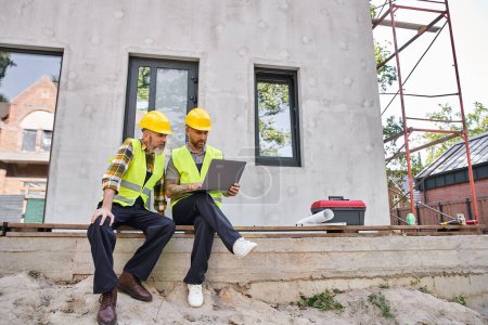 attrayants constructeurs de chalets barbus dans des casques de sécurité assis sur le porche et travaillant sur ordinateur portable