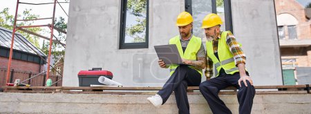 Photo for Appealing bearded cottage builders in safety helmets sitting on porch and working on laptop, banner - Royalty Free Image