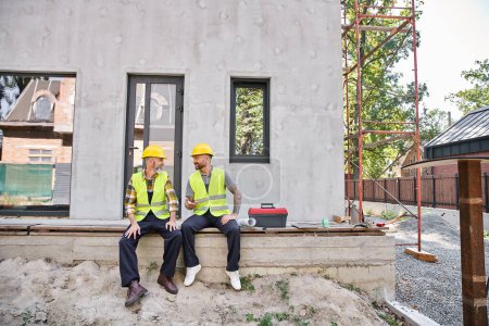 Photo for Joyful dedicated cottage builders in safety vests sitting on porch and looking at each other - Royalty Free Image