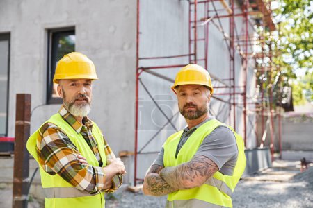Photo for Good looking cottage builders in safety helmets posing next to scaffolding and looking at camera - Royalty Free Image