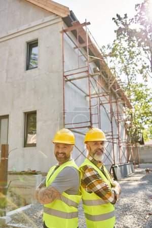 Photo for Cheerful cottage builders in safety vests posing back to back near scaffolding and looking at camera - Royalty Free Image