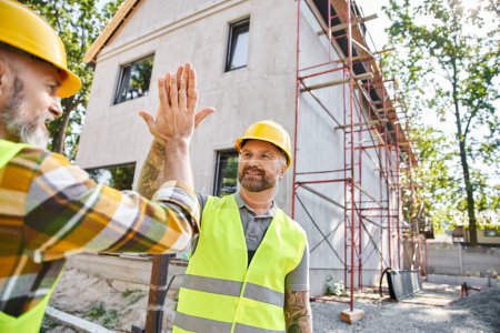 Photo for Two cheerful cottage builders in safety attires giving high five to each other near scaffolding - Royalty Free Image