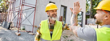 Photo for Two jolly cottage builders in safety attires giving high five to each other near scaffolding, banner - Royalty Free Image