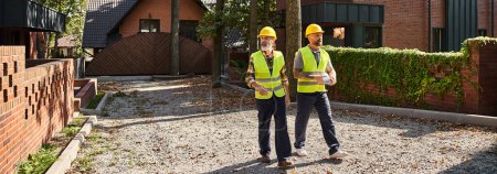 Photo for Handsome construction workers in safety vests walking and discussing site, cottage builders, banner - Royalty Free Image