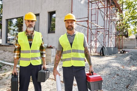 two attractive men in safety helmets and vests posing with toolboxes and blueprint, cottage builders