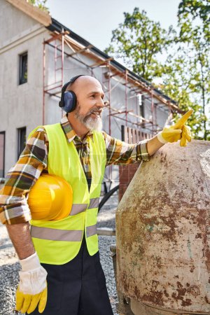 Foto de Alegre constructor de casa de campo en guantes de seguridad y chaleco posando con auriculares y casco en el sitio - Imagen libre de derechos