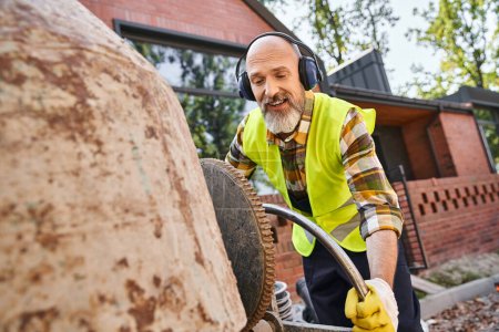 handsome cheerful man in safety gloves and headphones working with circular saw, cottage builder