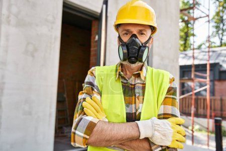 Foto de Atractivo hombre en guantes de seguridad y casco con máscara de polvo y mirando a la cámara, constructor de cabañas - Imagen libre de derechos