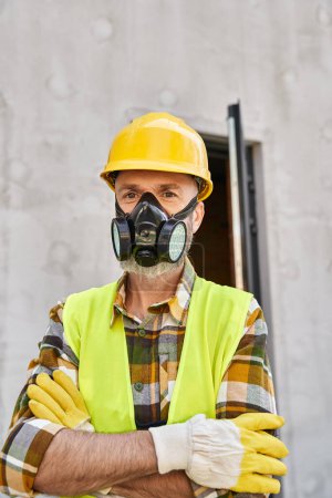 attractive man in safety gloves and helmet wearing dust mask and looking at camera, cottage builder