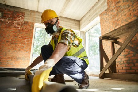 hardworking construction worker in safety helmet and gloves with dust mask putting carpet on floor