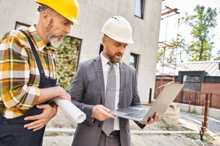 handsome architect and builder in overalls and suit working with laptop on construction site