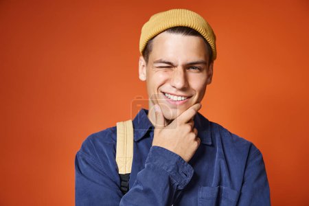 Porträt eines charmanten jungen Mannes mit gelbem Hut und grauen Augen, der zwinkert und das Kinn mit der Hand berührt