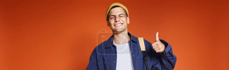 Téléchargez les photos : Bannière de jeune homme heureux en chapeau debout avec sac à dos et montrant comme sur fond de terre cuite - en image libre de droit