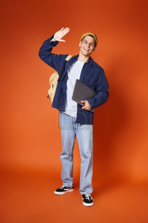 Photo for Young man in casual outfit and yellow hat with backpack holding laptop and saying hello - Royalty Free Image
