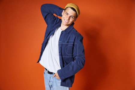cheerful young man in stylish outfit and yellow hat posing putting hand behind back