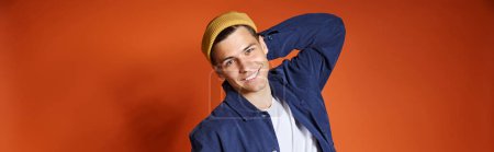 horizontal shot of young man in stylish outfit and yellow hat posing putting hand behind back