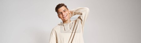 Photo for Banner of young man in white hoodie smiling and putting hand behind head against light background - Royalty Free Image