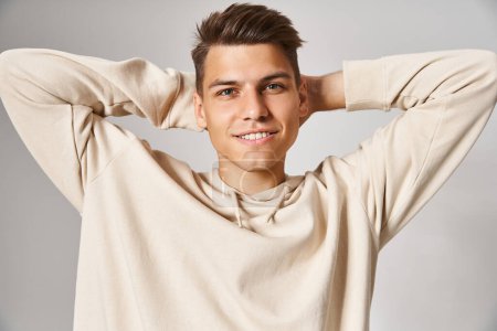 charismatic man in white hoodie smiling and putting hands behind head against light background