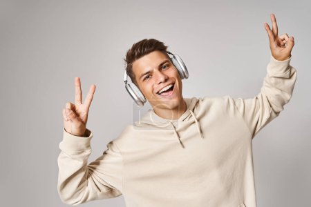 Photo for Cheerful student in headphones smiling and showing sign peace with hands against light background - Royalty Free Image