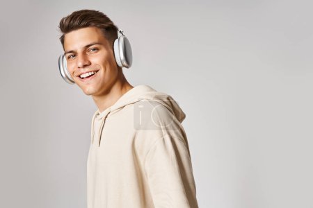 Téléchargez les photos : Attrayant jeune gars avec les cheveux bruns dans les écouteurs souriant à la caméra sur fond de lumière - en image libre de droit