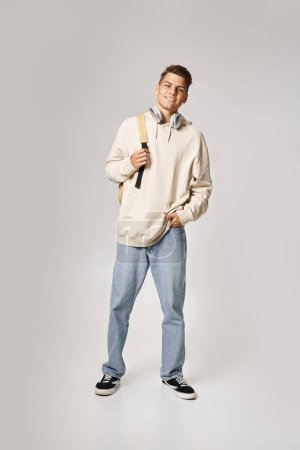 Foto de Estudiante guapo en auriculares y sudadera con capucha blanca con mochila contra fondo claro - Imagen libre de derechos