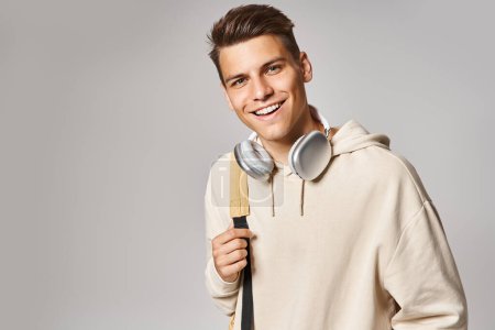 Foto de Atractivo joven estudiante en auriculares y atuendo casual con mochila sobre fondo gris - Imagen libre de derechos