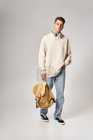 Foto de Estudiante guapo en auriculares y ropa casual caminando con mochila contra fondo gris - Imagen libre de derechos