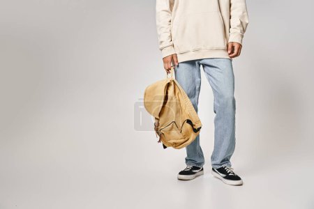 Schnappschuss eines Studenten in lässigem Outfit, der mit Rucksack vor grauem Hintergrund steht