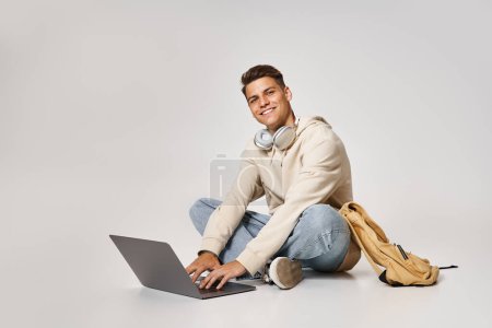 Téléchargez les photos : Gars gai dans le réseau casque pour ordinateur portable et assis avec sac à dos sur fond gris - en image libre de droit