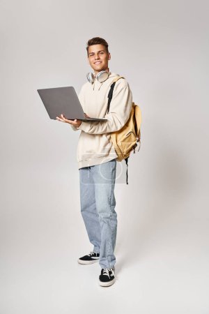 Téléchargez les photos : Jeune étudiant dans un casque debout avec sac à dos et réseautage pour ordinateur portable sur fond gris - en image libre de droit
