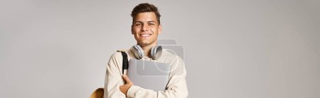 Téléchargez les photos : Plan horizontal de l'élève dans les années 20 avec ordinateur portable et écouteurs confiants debout sur fond gris - en image libre de droit