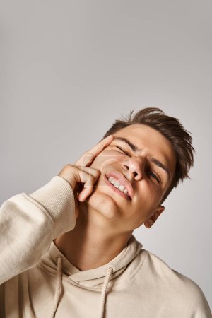 Porträt eines jungen Mannes mit braunen Haaren machen schiefe Gesicht mit Hand vor grauem Hintergrund
