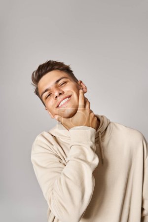 attractive man in his 20s with brown hair touching to jawline with hand on grey background