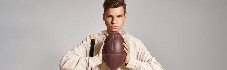 Téléchargez les photos : Plan horizontal de l'élève confiant avec sac à dos tenant la balle de rugby avec les mains sur fond gris - en image libre de droit