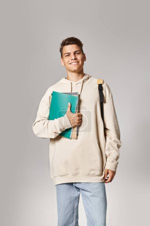 fröhlicher Student in seinen Zwanzigern mit Rucksack und Geldscheinen vor grauem Hintergrund