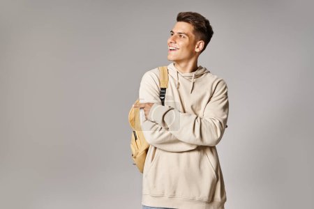 Foto de Atractivo joven con mochila y brazos cruzados mostrando el lado con el dedo sobre fondo gris - Imagen libre de derechos